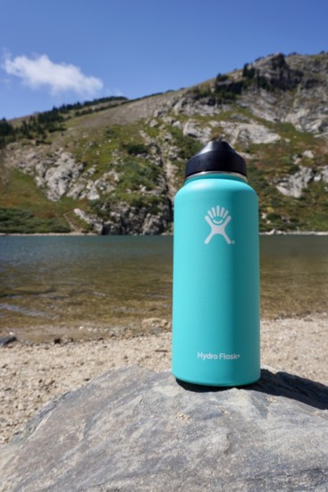 hydroflask bottle sitting on a rock with a lake and mountain in the background, offering a gift idea for hikers