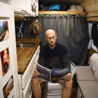 Jake sitting in the van on the bathroom toilet reading a magazine