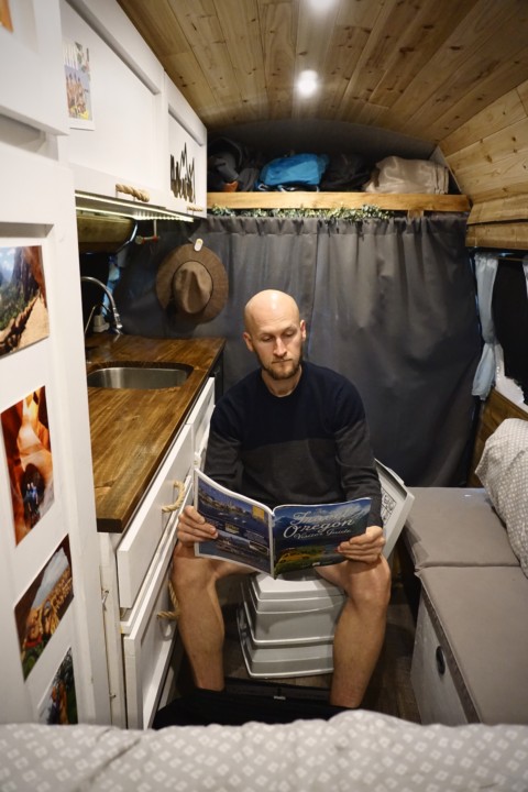 Jake sitting in the van on the bathroom toilet reading a magazine