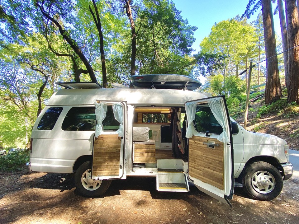 Two Roaming Souls Van with doors open showing the inside under bright green trees