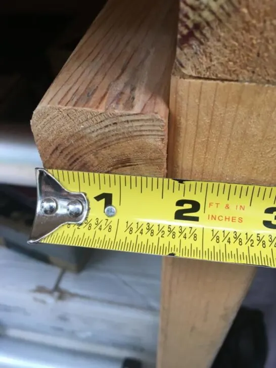 A "2x2" piece of wood with a measuring tape showing 1 1/2 inches. Understanding nominal vs actual dimensions is a crucial woodworking tip for van conversions.