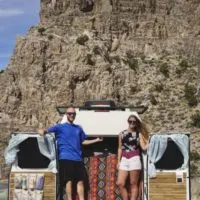 Jake and Emily standing on the rear bumper of their van with the doors open with a rock wall behind them