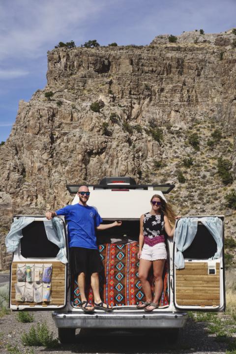 Jake and Emily standing on the rear bumper of their van with the doors open with a rock wall behind them