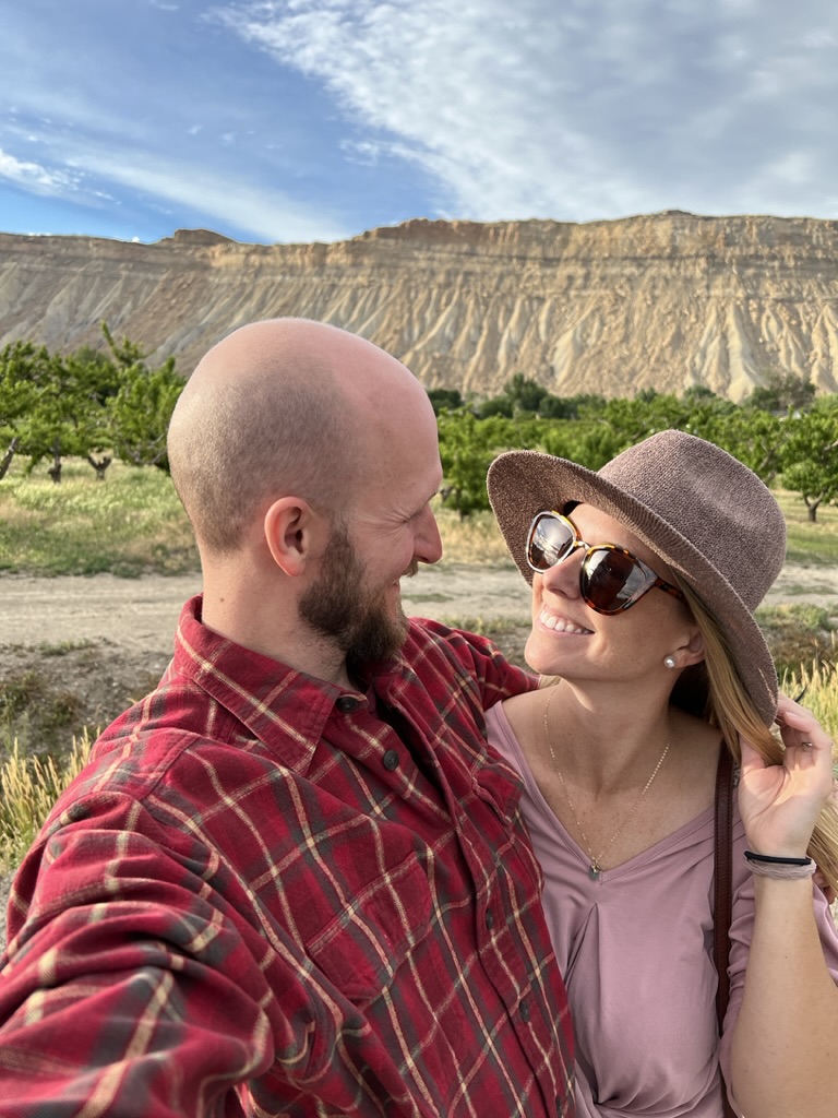 a couple looking and smiling at each other at a Harvest Hosts Location with a vineyard and cliff edge in the background