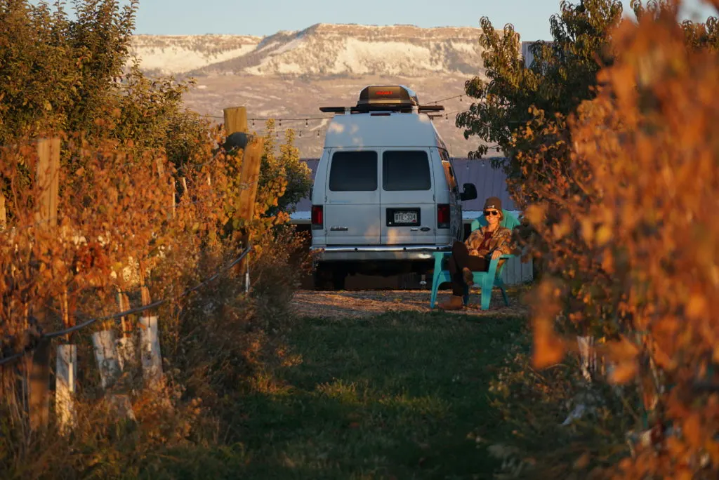 emily sitting in an adirondack chair in a vinyard with the vines, a campervan, and mountains in the background which is showing a Harvest Hosts