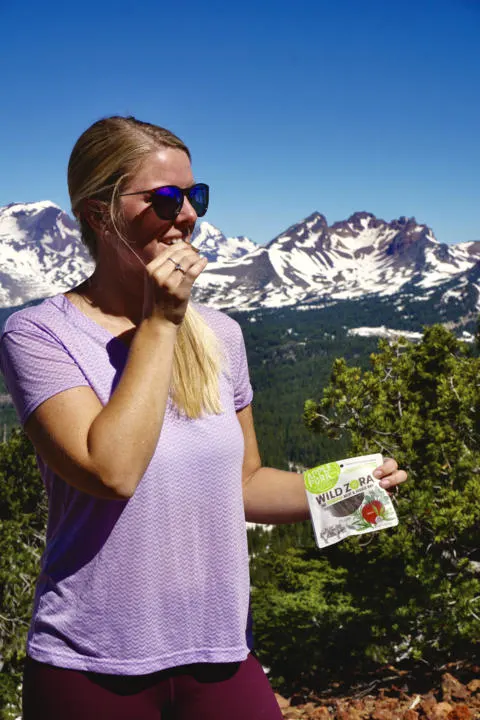 Emily eating a Wild Zora Meat & Veggie Bar With the Tumalo Mountains in the background
