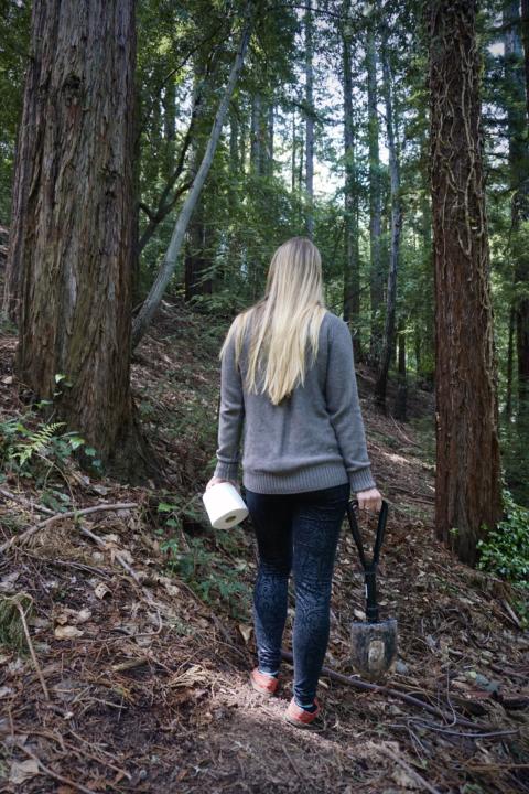 Emily walking into the woods with toilet paper and a shovel to show to show how to poop in the woods which is an off-grid bathroom option