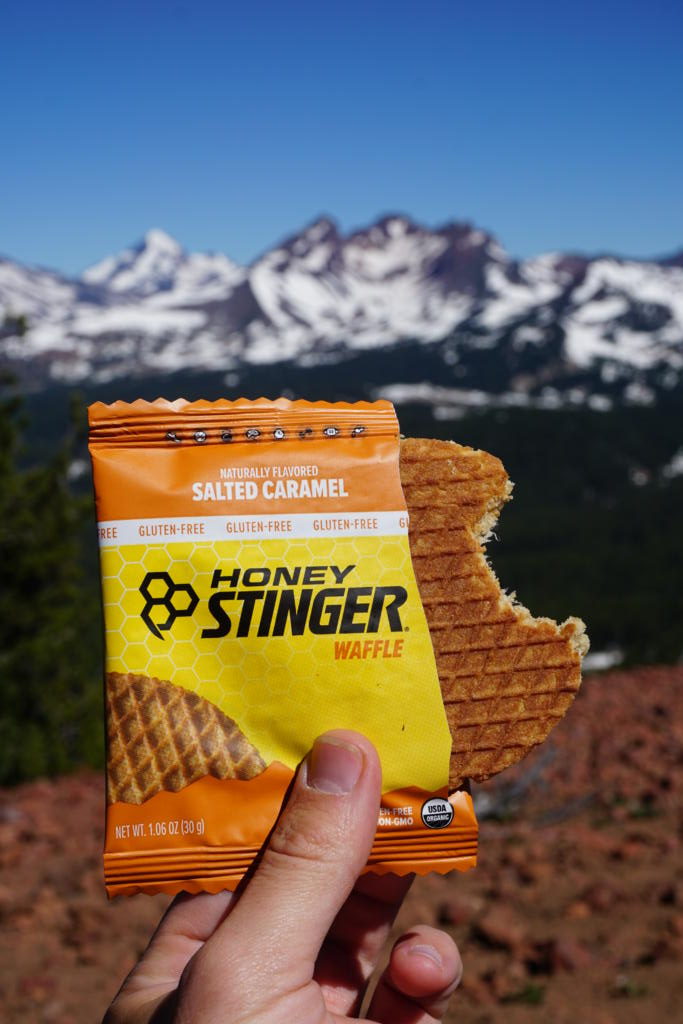 a bite out of a honey stinger waffle with mountains in the background which is one of the best hiking snacks to fuel your next adventure