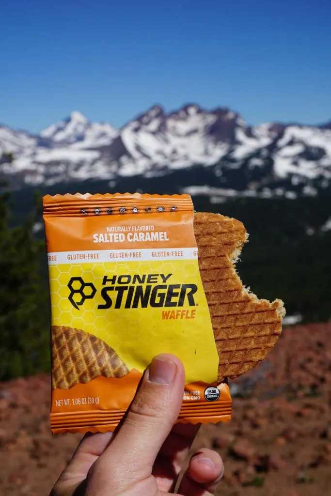 a bite out of a honey stinger waffle with mountains in the background which is one of the best hiking snacks to fuel your next adventure