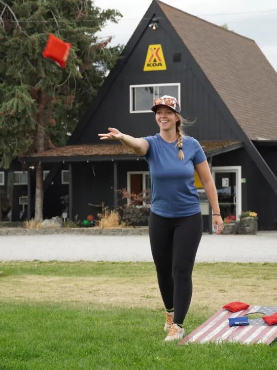 Emily throwing a bag at the cornhole board with the KOA building in the background